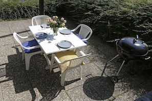 An outdoor dining area with a white table set for four, surrounded by white chairs, a vase of flowers, and a barbecue grill.