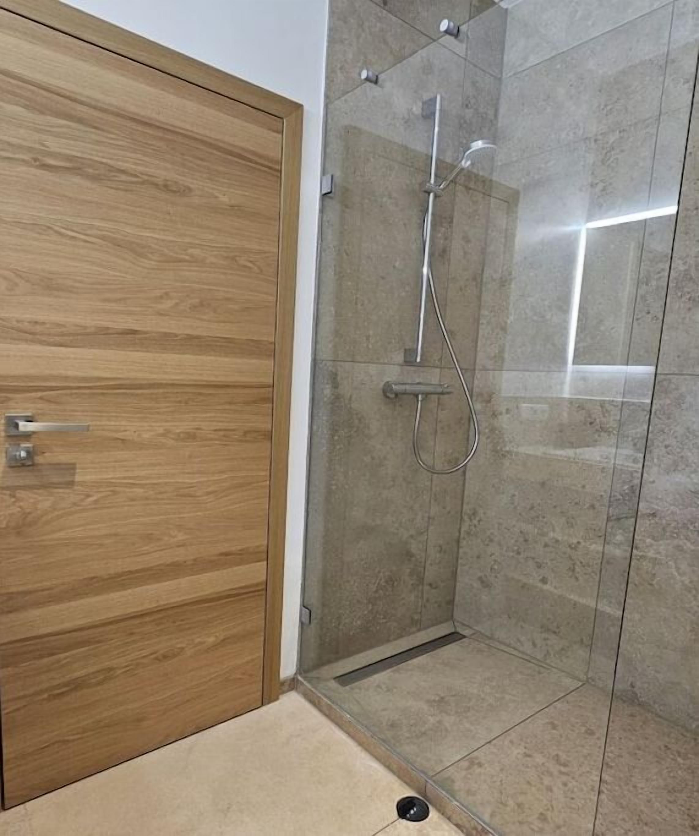 A shower with a glass door, a wooden door to the left, and a tiled floor.