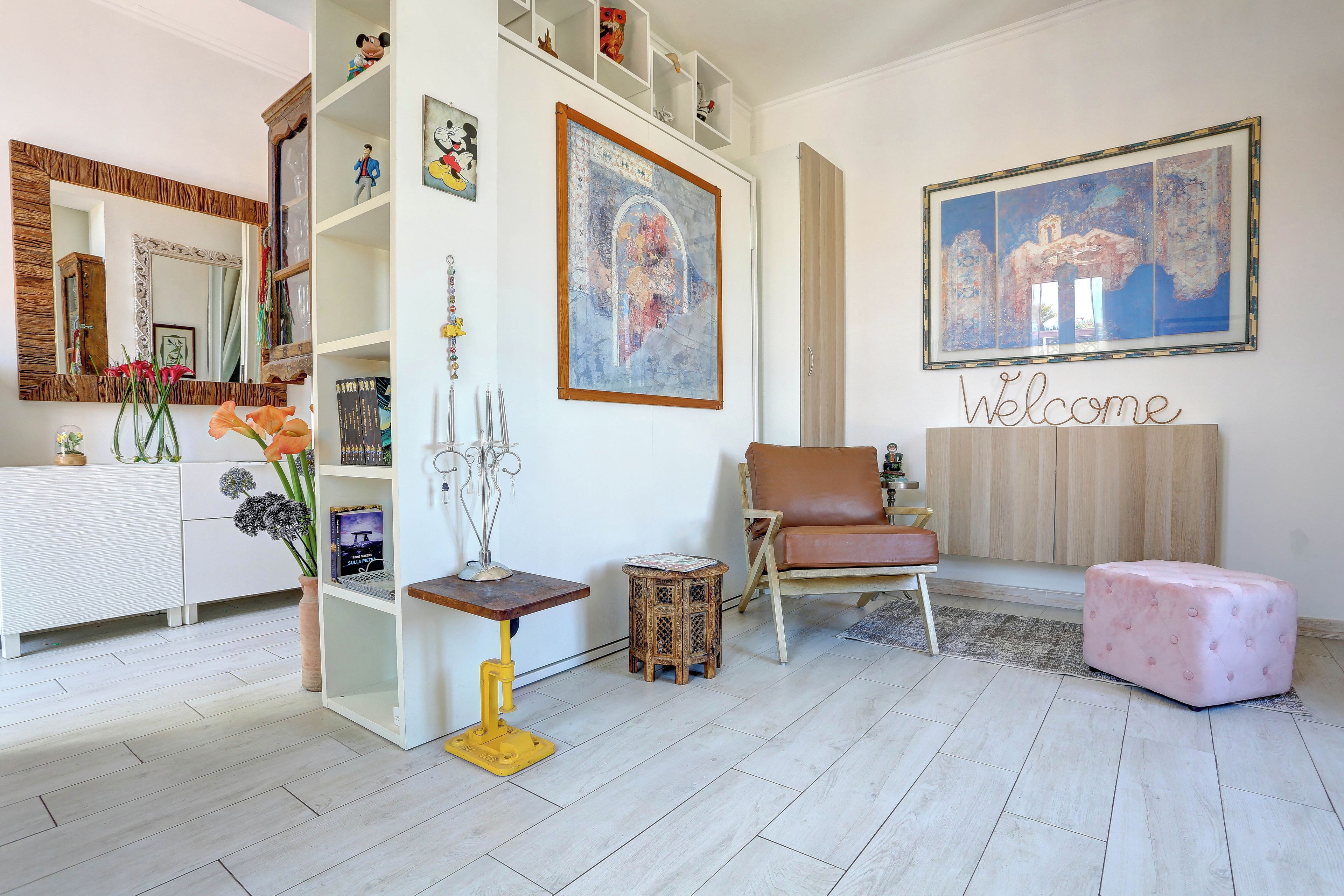 Una sala de estar moderna con un sofá, una silla, una mesita y un otoman rosa. Se observa una estantería con diversos objetos y un cuadro grande en la pared.