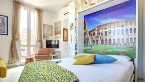 Et hotelværelse med seng, fjernsyn, skrivebord og et stort vindue med udsigt til Colosseum.