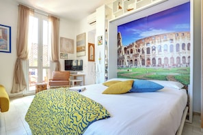 Habitación de hotel con cama, televisor, escritorio y una ventana grande con vistas al Coliseo.
