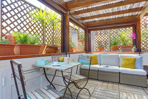 An outdoor patio with a wooden table, wicker sofa, and potted plants. - COLOSSEO ROOFTOP 36 - Exclusive Terrace, Solarium & Skyline View (Roma)