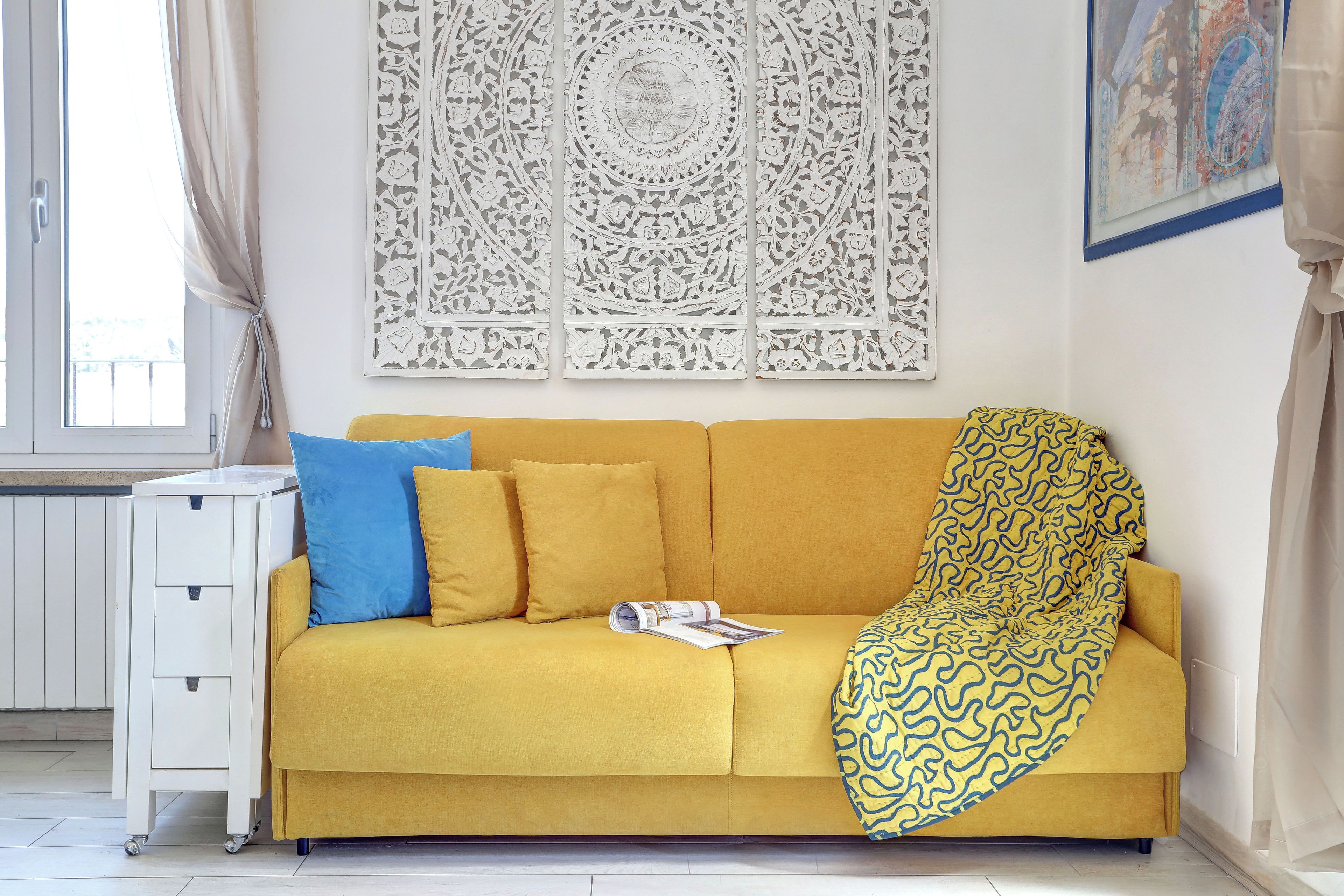 A yellow sofa with a patterned throw blanket, a blue pillow, and a white side table with a magazine.