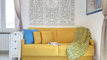 Un sofá amarillo con una manta de diseño, una almohada azul y una mesita auxiliar blanca con una revista.