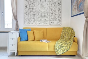 Un sofá amarillo con una manta de diseño, una almohada azul y una mesita auxiliar blanca con una revista.