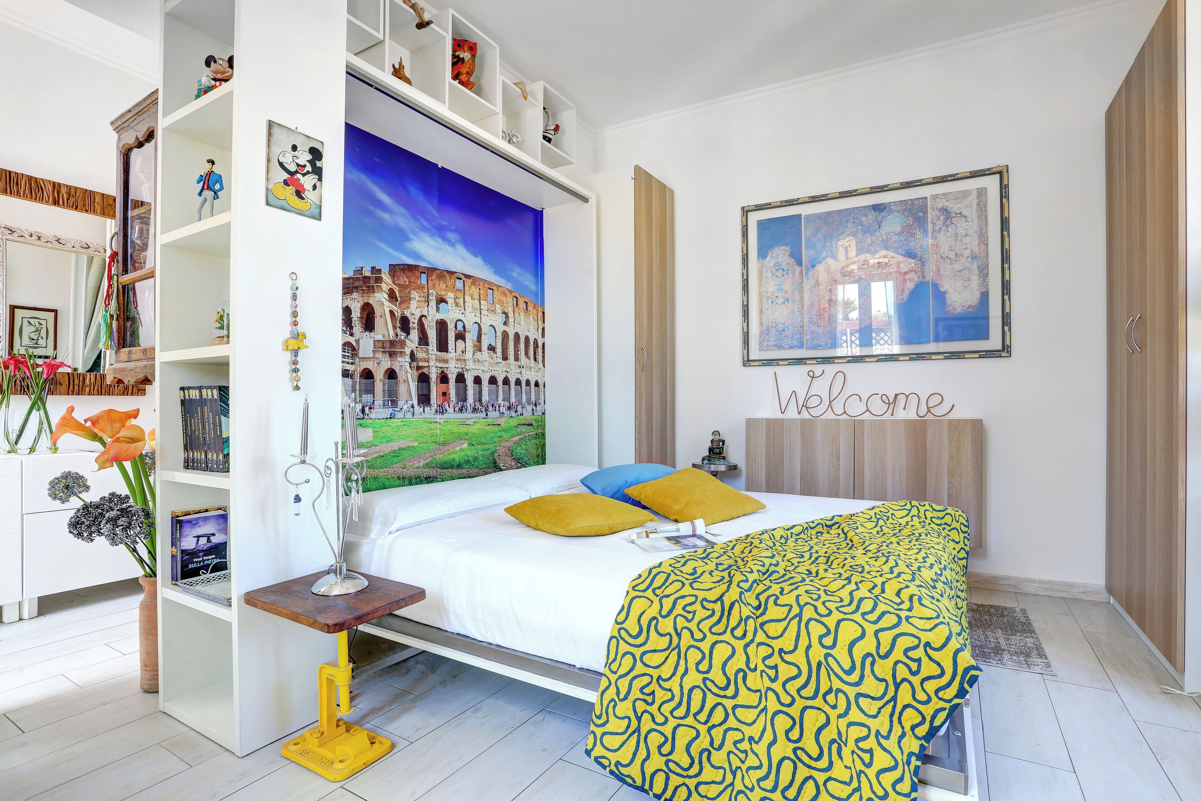 Un dormitorio con una cama, una estantería, un escritorio y una pared con un cartel de bienvenida.