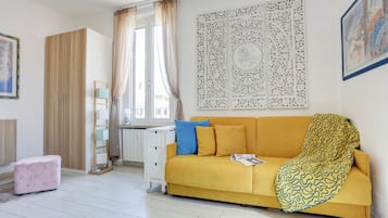 Una habitación con sofá amarillo, reposapiés rosa, escritorio blanco y ventana con cortinas.