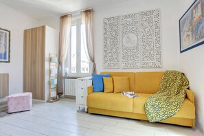 Una habitación con sofá amarillo, reposapiés rosa, escritorio blanco y ventana con cortinas.