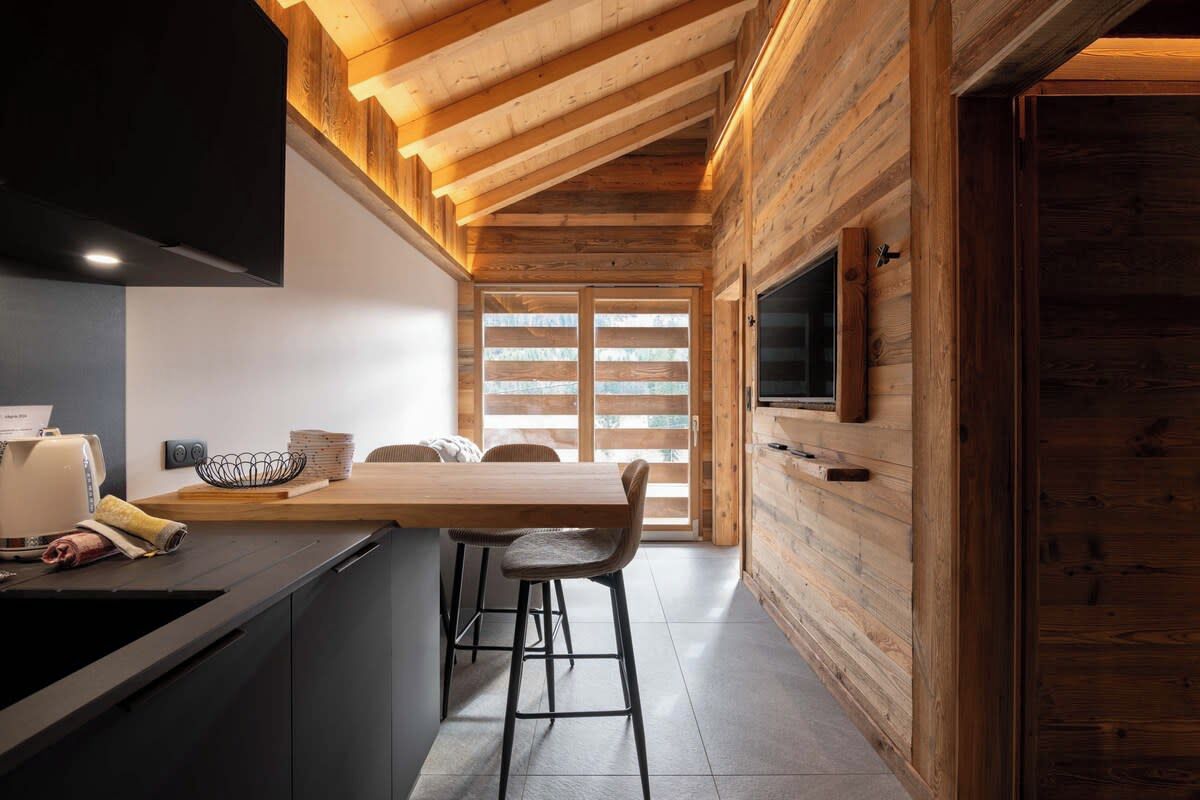 A modern kitchen with a wooden ceiling, a dining area with a table and chairs, and a TV mounted on the wall.