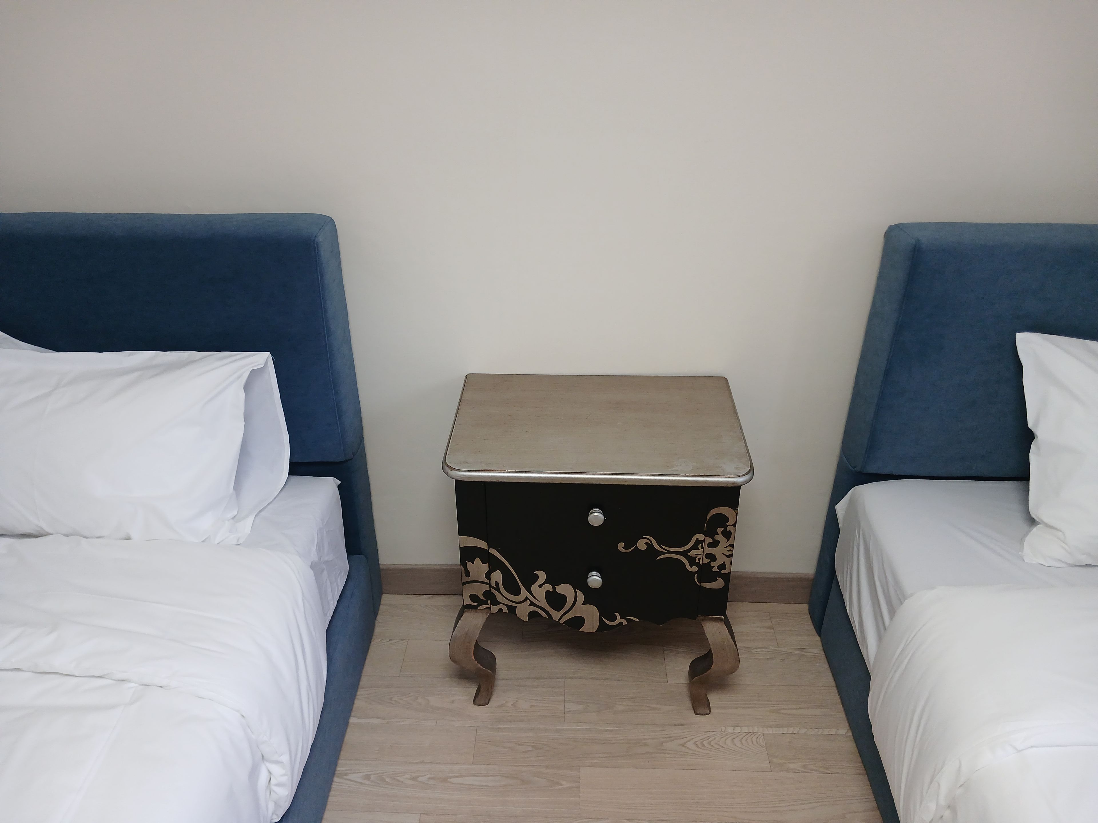 A room with two beds, each with white bedding, and a small bedside table with a decorative design.