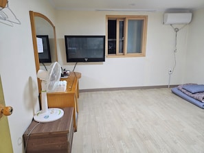A room with a flat-screen TV, a wooden desk, a standing fan, a heater, a window with blinds, and a bed.