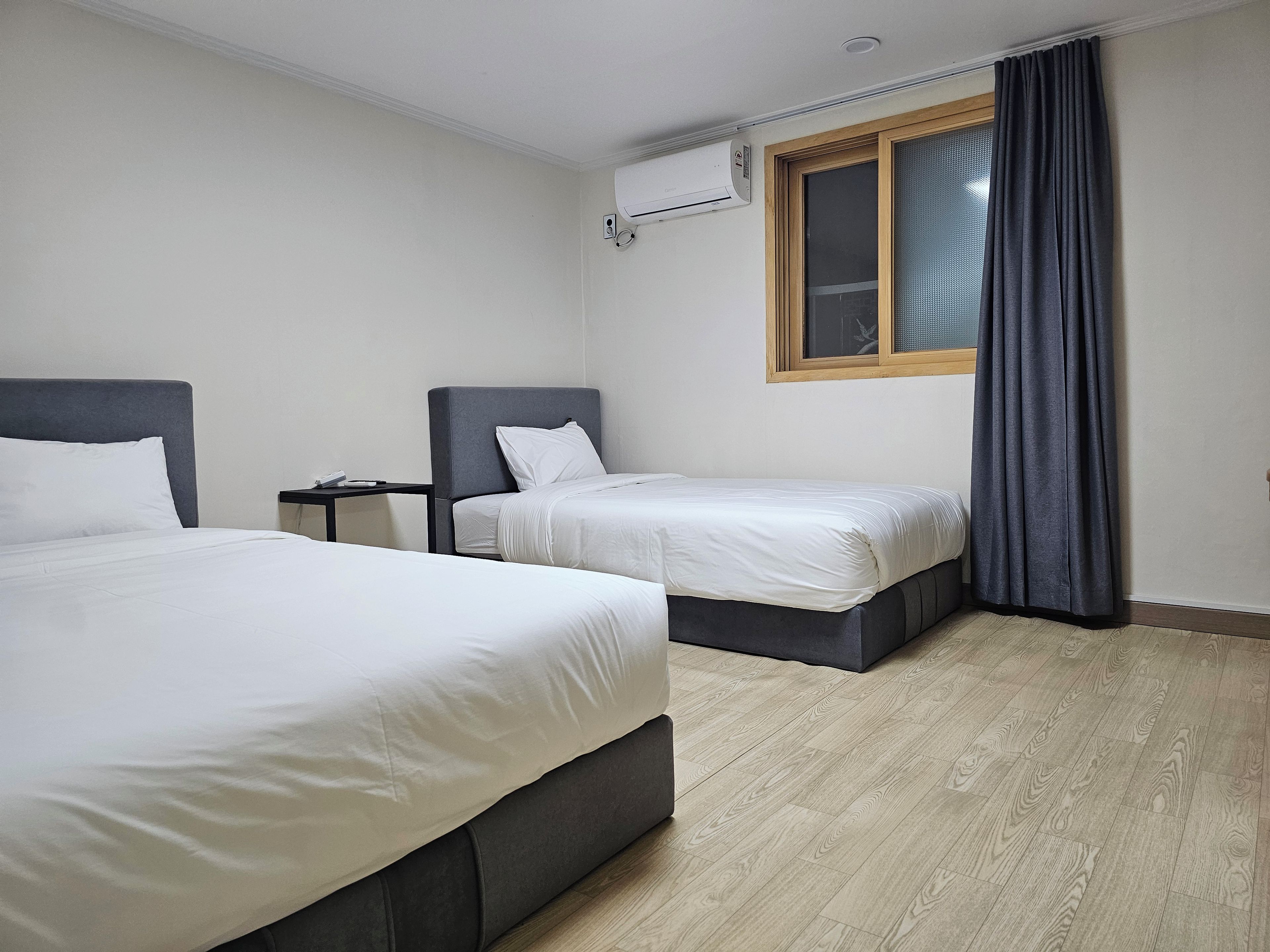 Habitació d'hotel amb dos llits, una petita taula de nit, un aparell d'aire condicionat, una finestra amb cortines i un sòl de fusta.