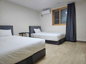 Habitación de hotel con dos camas, una mesita de noche pequeña, un equipo de aire acondicionado, una ventana con cortinas y piso de madera.