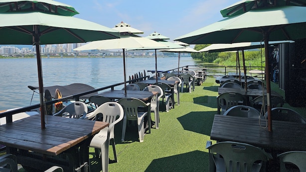Tavoli per mangiare all'aperto con ombrelloni, sedie e vista su un fiume con lo skyline di una città sullo sfondo.