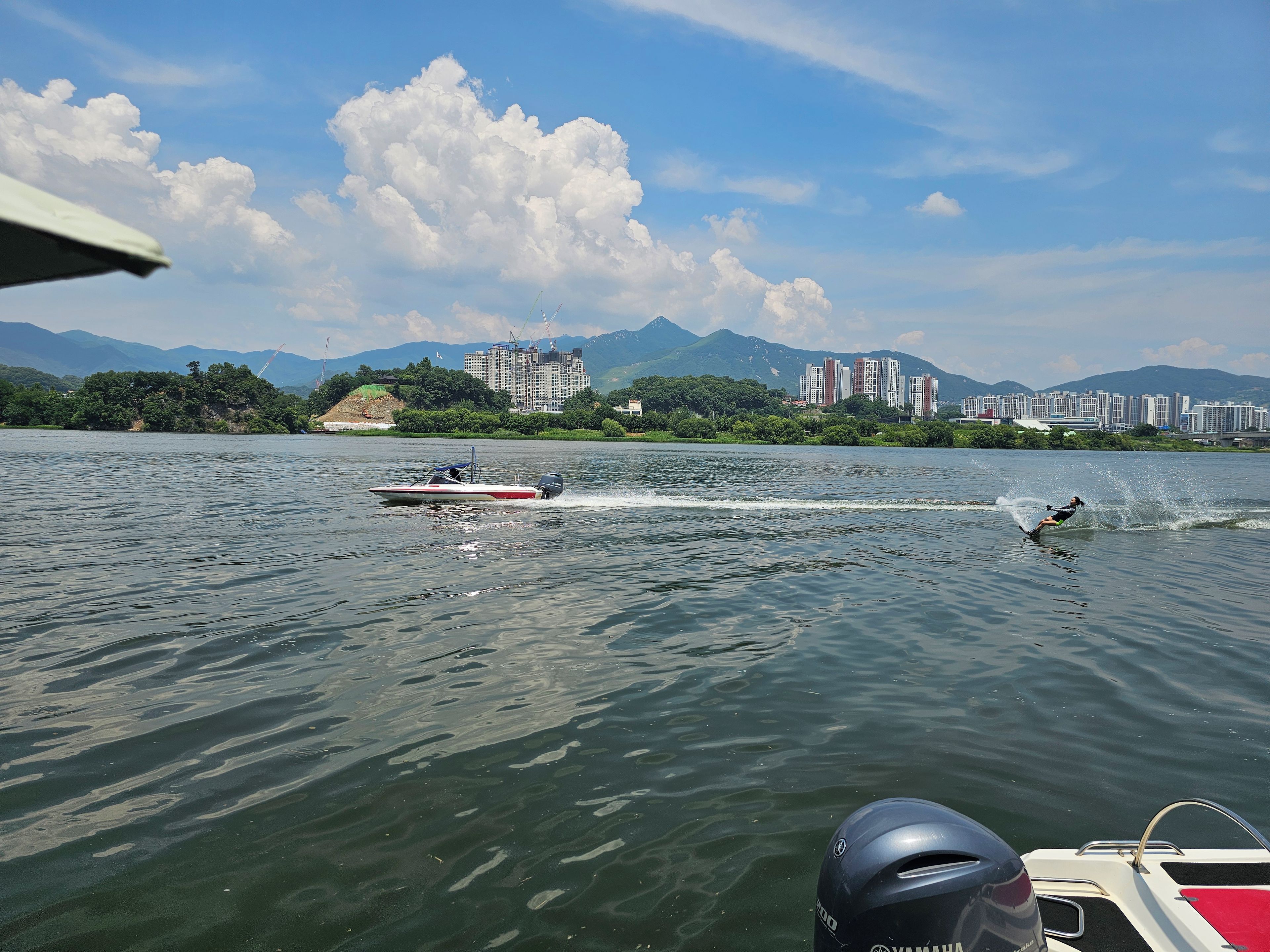 A body of water with a speedboat and a wakeboarder. In the background, there are buildings and mountains.