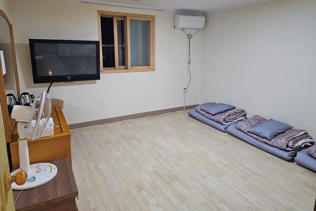 A room with a flat-screen TV mounted on the wall, a standing fan, a heater, a window with blinds, and a sofa with blankets.
