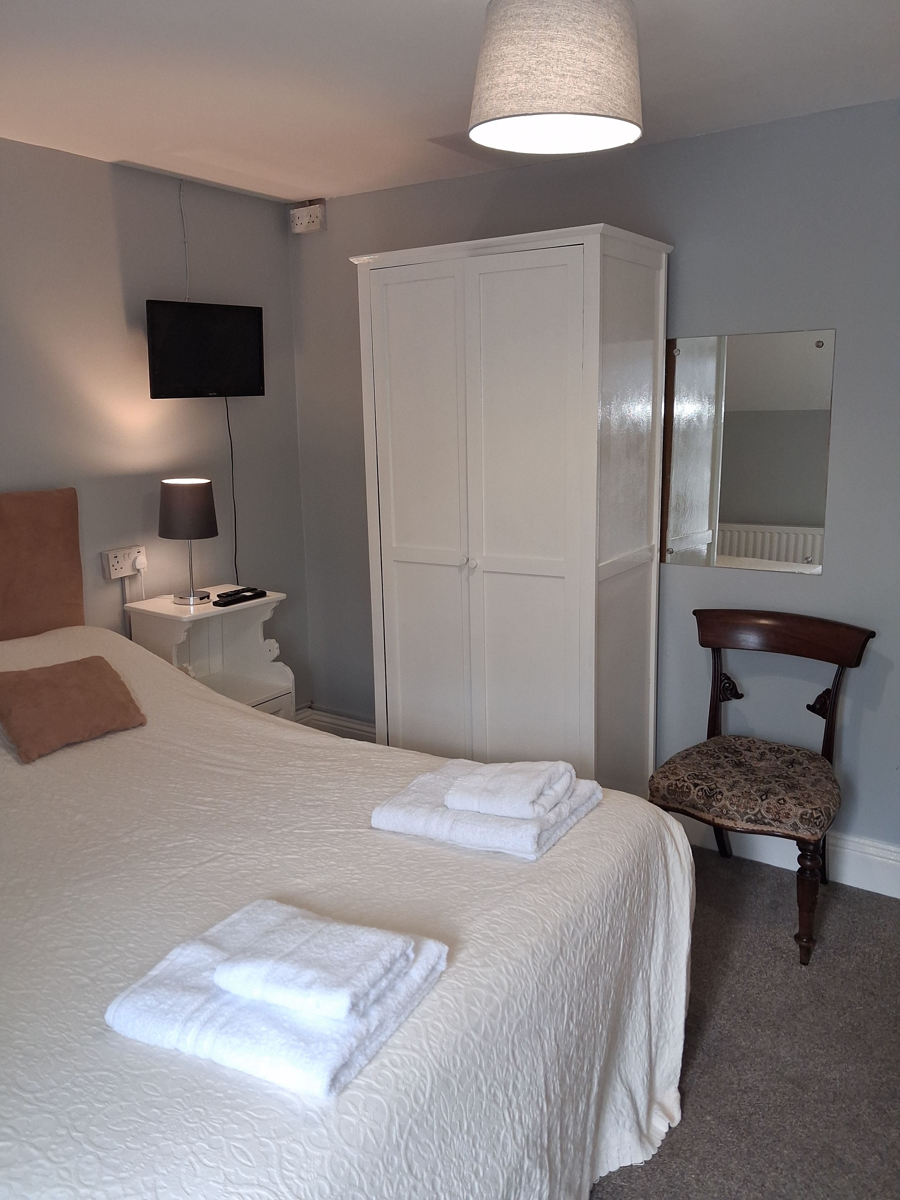Een slaapkamer met een bed, een kledingkast, een stoel, een nachtkastje, een lamp, een televisie en een spiegel.