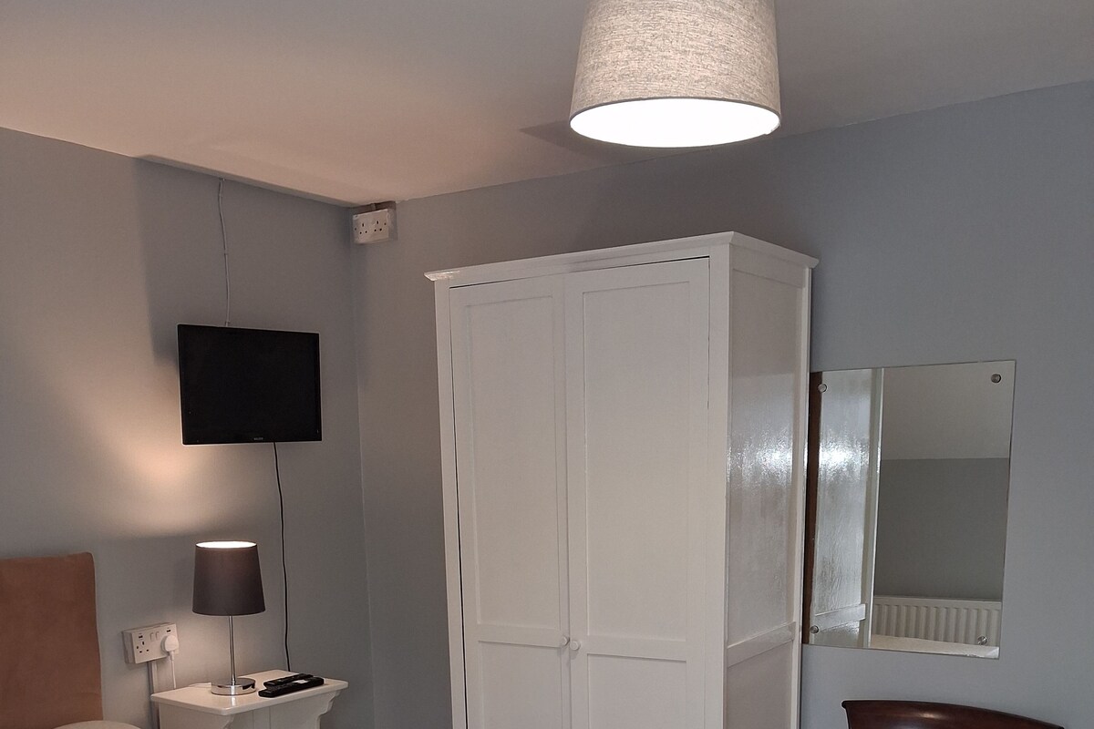 Un dormitorio con una cama, una mesita de noche con lámpara, un armario, una silla y un televisor en la pared.