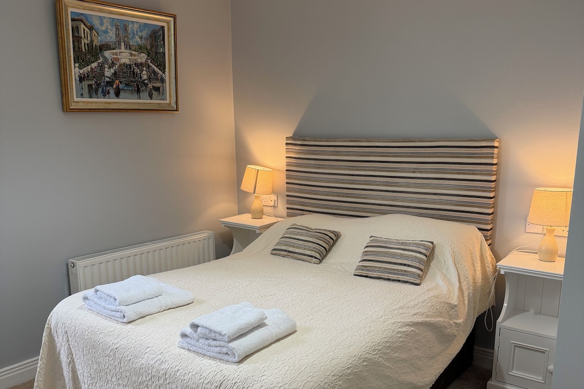 Una cama bien tendida con sábanas blancas y toallas dobladas, cabecera con rayas horizontales, lámparas de noche y un cuadro en la pared.