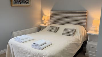 A neatly made bed with white linens and folded towels, a headboard with horizontal stripes, bedside lamps, and a framed painting on the wall.
