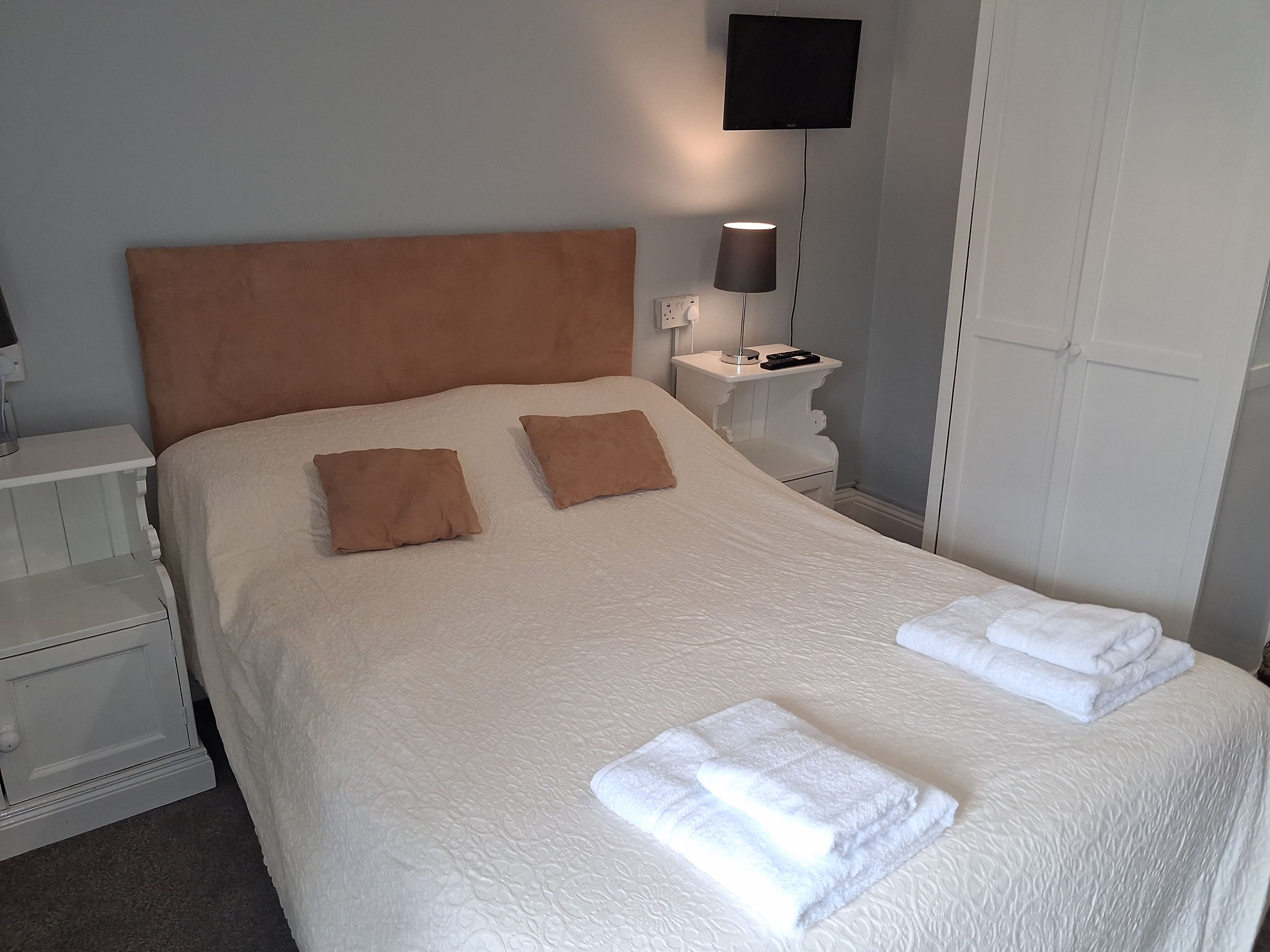 Un lit bien fait, avec deux oreillers et deux serviettes pliées, une lampe de chevet et un téléviseur à écran plat fixé au mur.