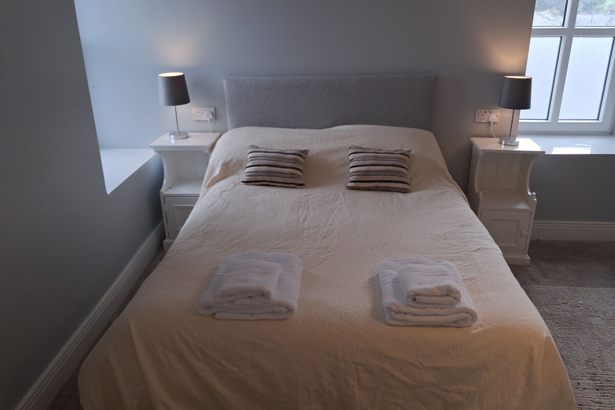 Una cama bien hecha con dos almohadas, dos lámparas de noche y dos toallas dobladas.