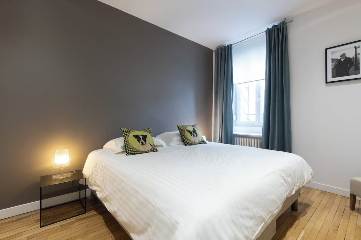 Une chambre avec un grand lit, une table de chevet avec une lampe, une fenêtre avec des rideaux et un tableau encadré au mur.