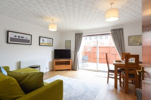 Ett vardagsrum med en soffa, en tv, ett matbord och stolar. Det finns ett fönster med gardiner och utsikt över omgivningen.
