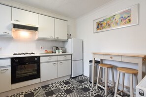 Cocina compacta con alacenas blancas, estufa a gas, refrigerador y máquina de café. El piso presenta un diseño y hay un cuadro en la pared.