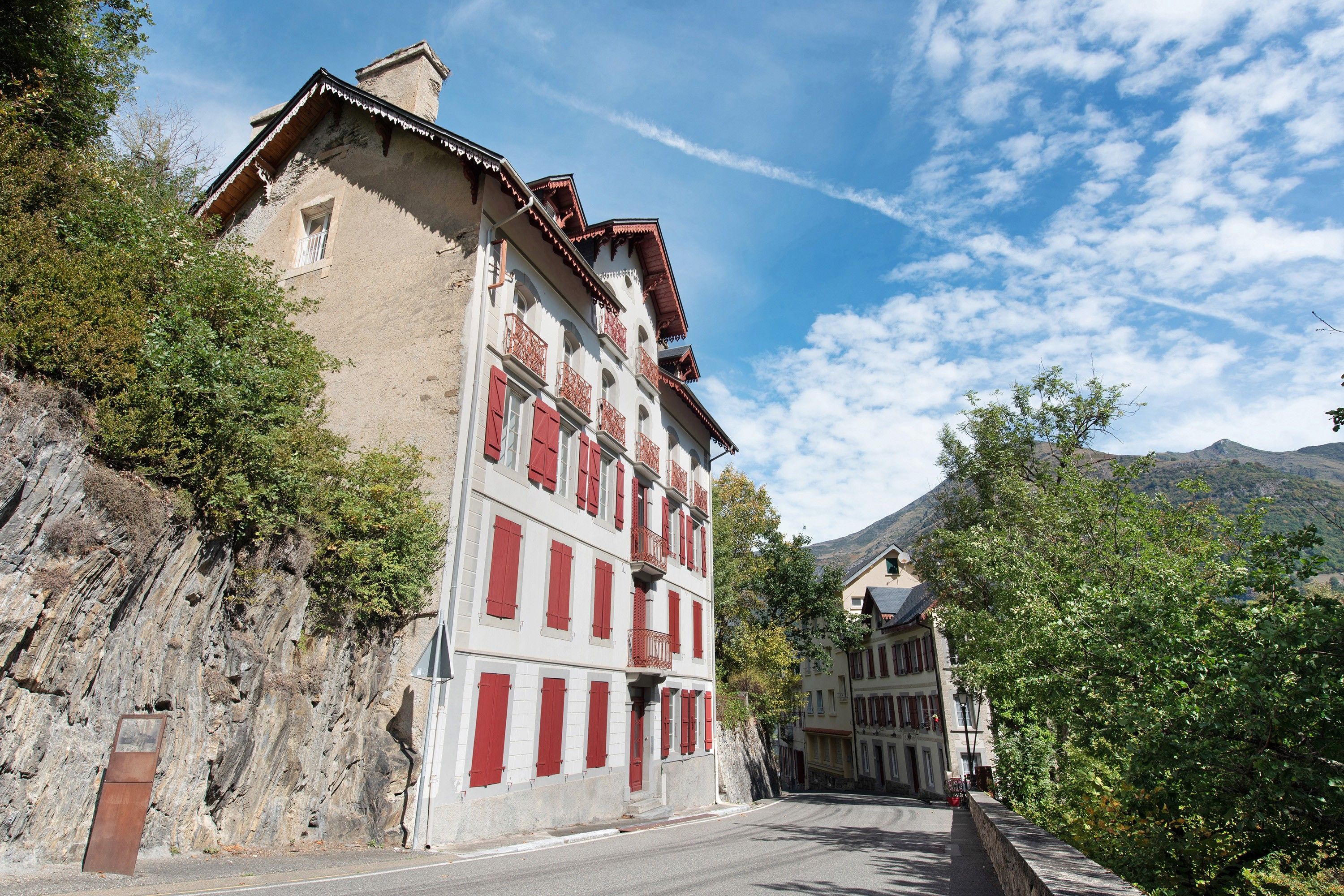 A street with buildings, one with red shutters and balconies, and a mountain in the background.