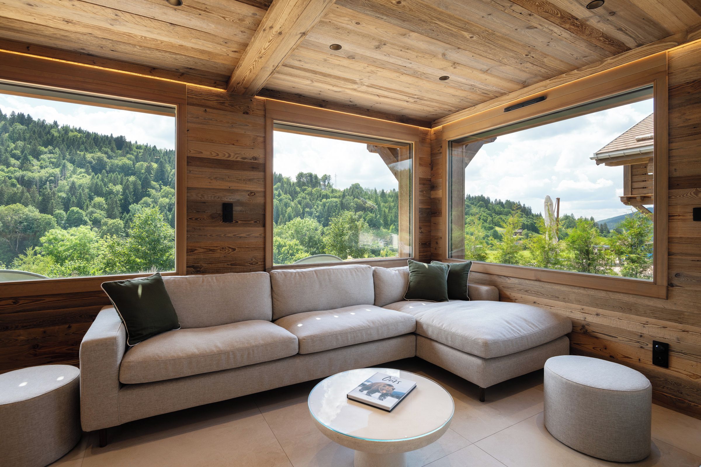 A modern interior with a large L-shaped sectional sofa, a glass coffee table, and a view of a forested landscape through large windows.