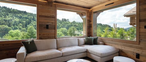 A modern interior with a large L-shaped sectional sofa, a glass coffee table, and a view of a forested landscape through large windows.
