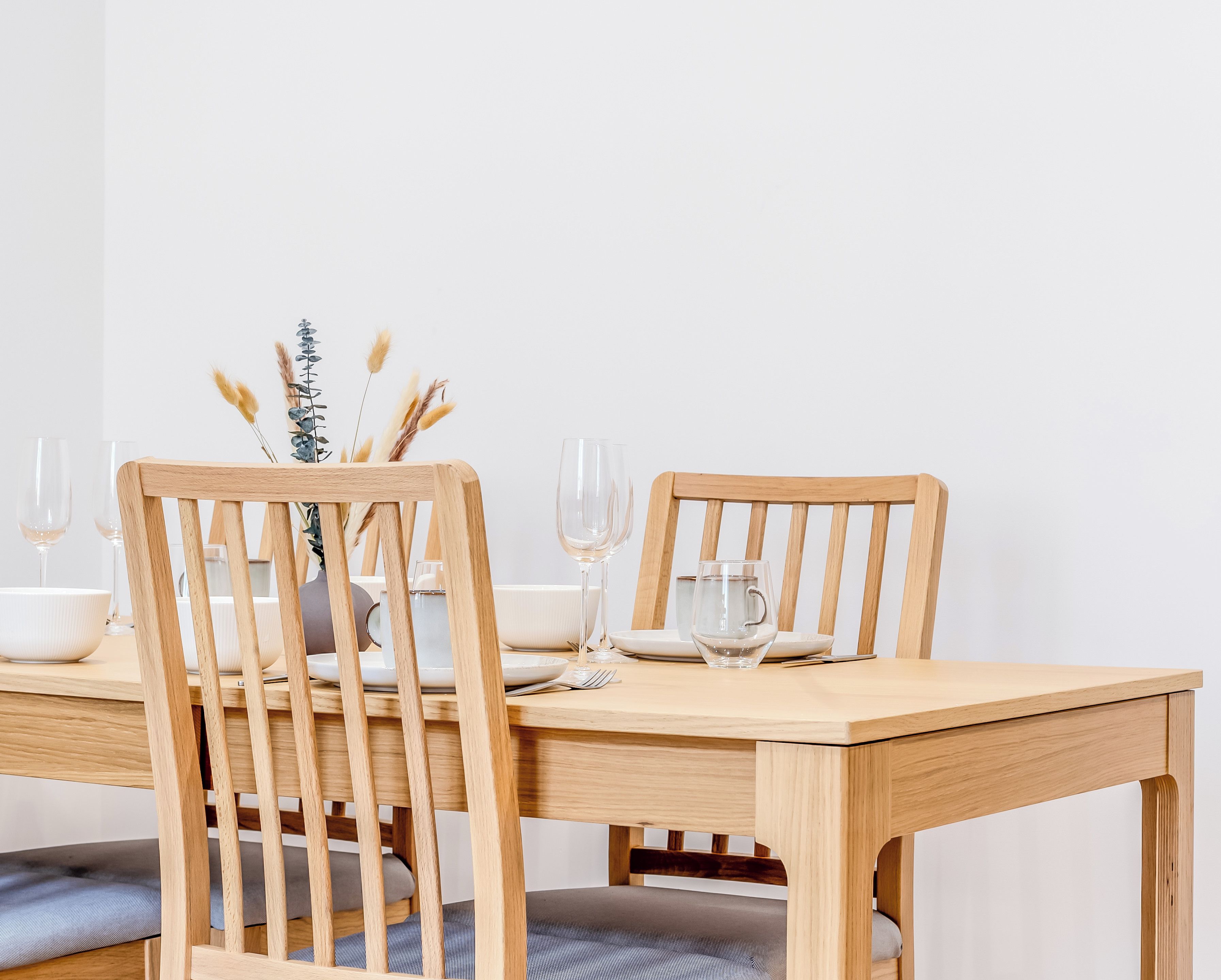 Een houten eettafel met twee stoelen, een vaas met gedroogde planten en lege wijnglazen.
