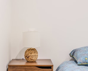 Ein Nachttisch mit einer Lampenschirm und einem gewobenen Fuß, einer Holzkassette sowie ein Bett mit einem blau-gelb gemusterten Kissen.