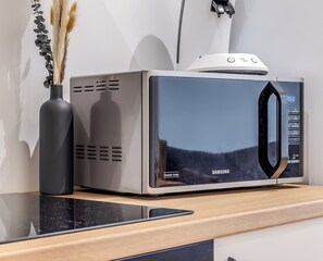 Eine Samsung-Mikrowelle mit digitaler Zeitanzeige, auf einer Holzauslage neben einer schwarzen Vase mit Trockenpflanzen platziert.