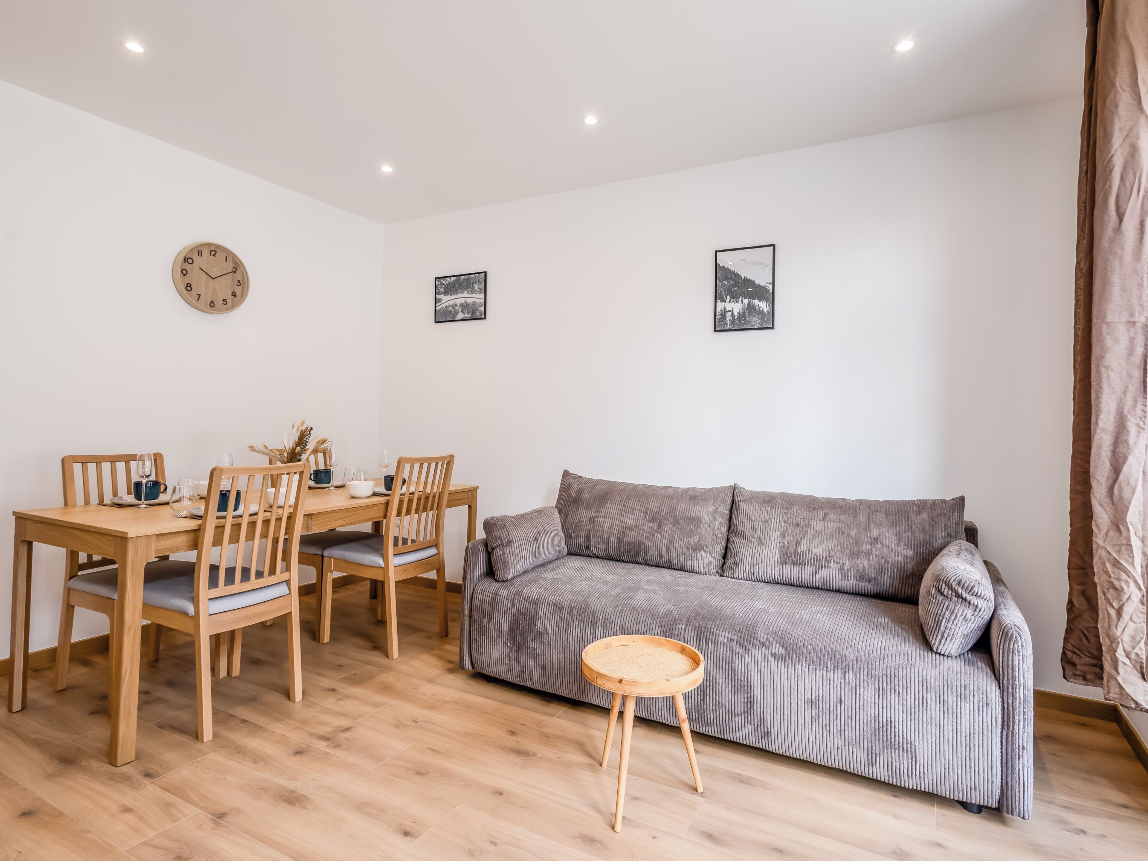 Een kamer met een houten eettafel, stoelen, een grijze bank, een kleine ronde tafel en twee ingelijste foto's aan de muur.