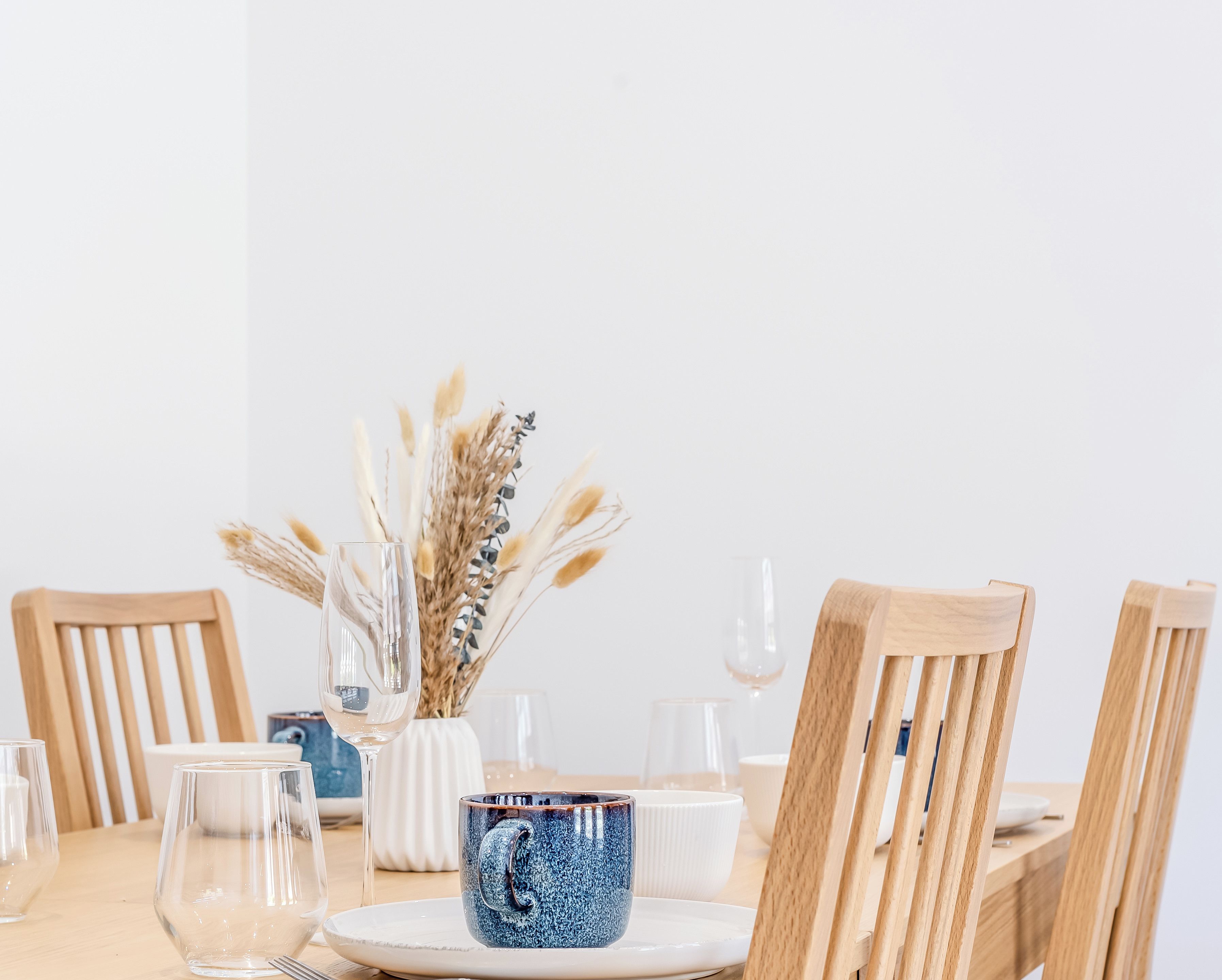 Ein Esstisch ist gedeckt mit klarem Glas, einer Keramiktasse und einer Vase mit getrockneten Pflanzen.