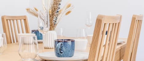 Ein Esstisch ist gedeckt mit klarem Glas, einer Keramiktasse und einer Vase mit getrockneten Pflanzen.