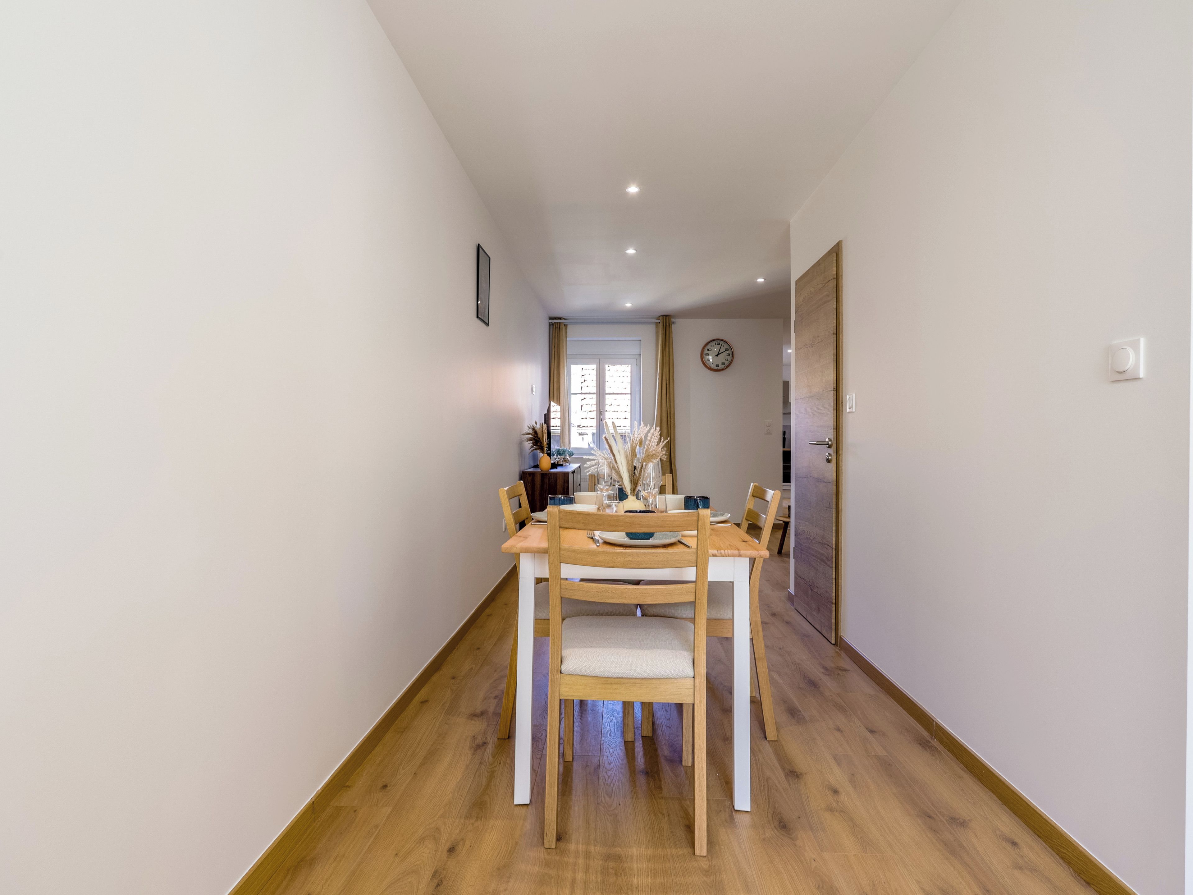 Een eetruimte met een houten tafel en stoelen, een klok aan de muur, en een deur die naar een andere kamer leidt.