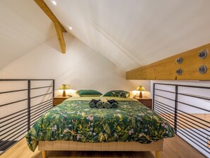 Ein Schlafzimmer mit schrägen Wänden, einem Bett mit grünem Blumenüberwurf, zwei Nachttischen mit Lampen und einem Holzbalken über dem Bett.