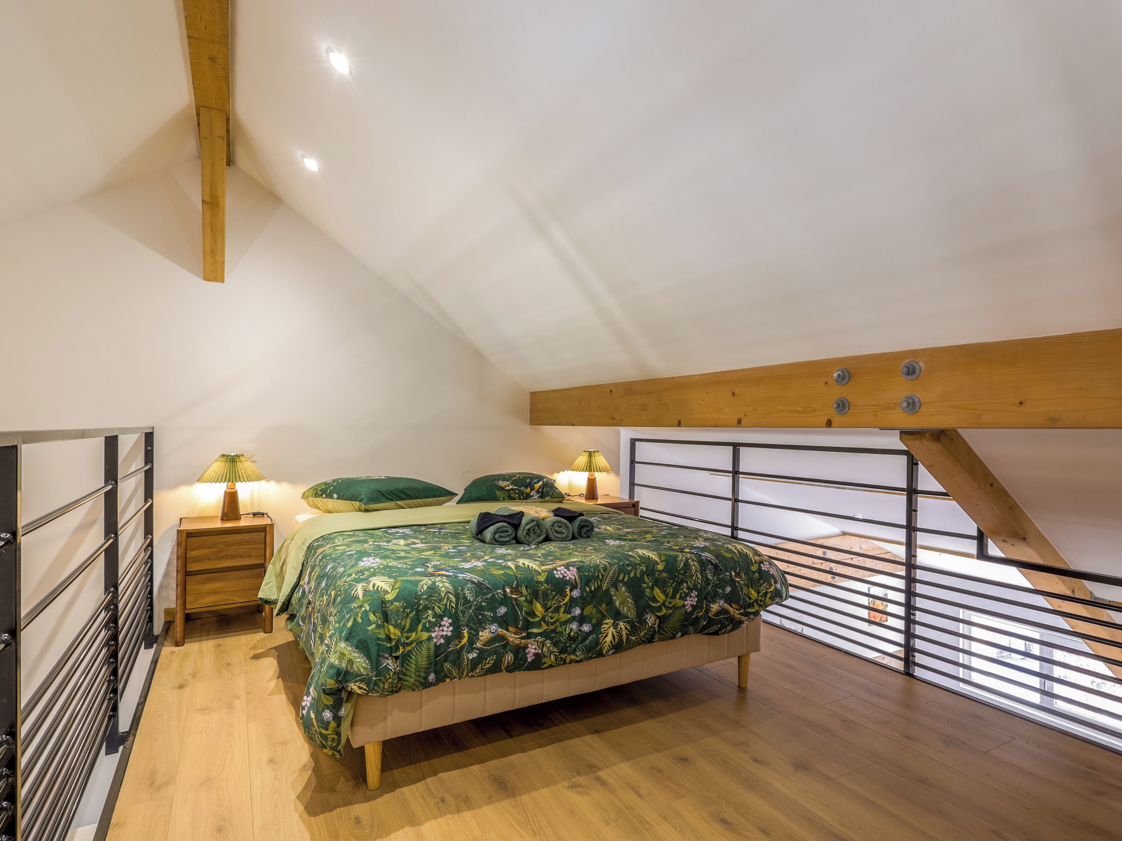 Ein Schlafzimmer mit schrägem Dach, einem Bett mit blumigem Überwurf, zwei Nachttischlampen und Blick ins Freie durch ein Fenster.