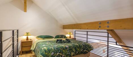 Ein Schlafzimmer mit schrägem Dach, einem Bett mit blumigem Überwurf, zwei Nachttischlampen und Blick ins Freie durch ein Fenster.