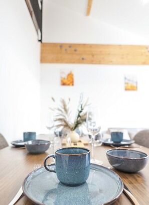 Een eettafel met een blauwe keramische mok, een bord en een kom. Achtergrond is een vaas met gedroogde planten en twee kleine blauwe bekers.