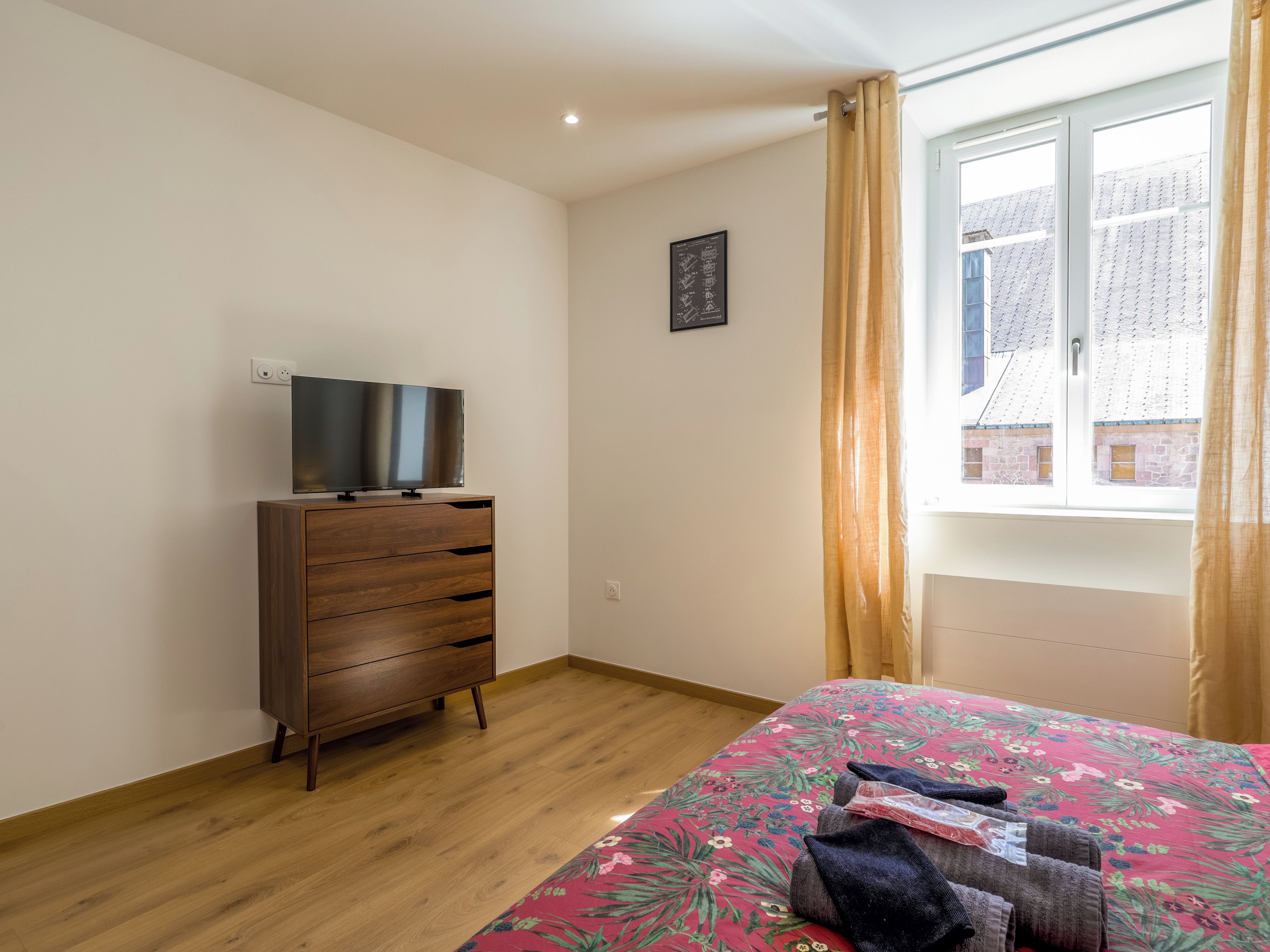 A bedroom with a wooden dresser, a flat-screen TV, a floral bedspread, and a window with curtains.