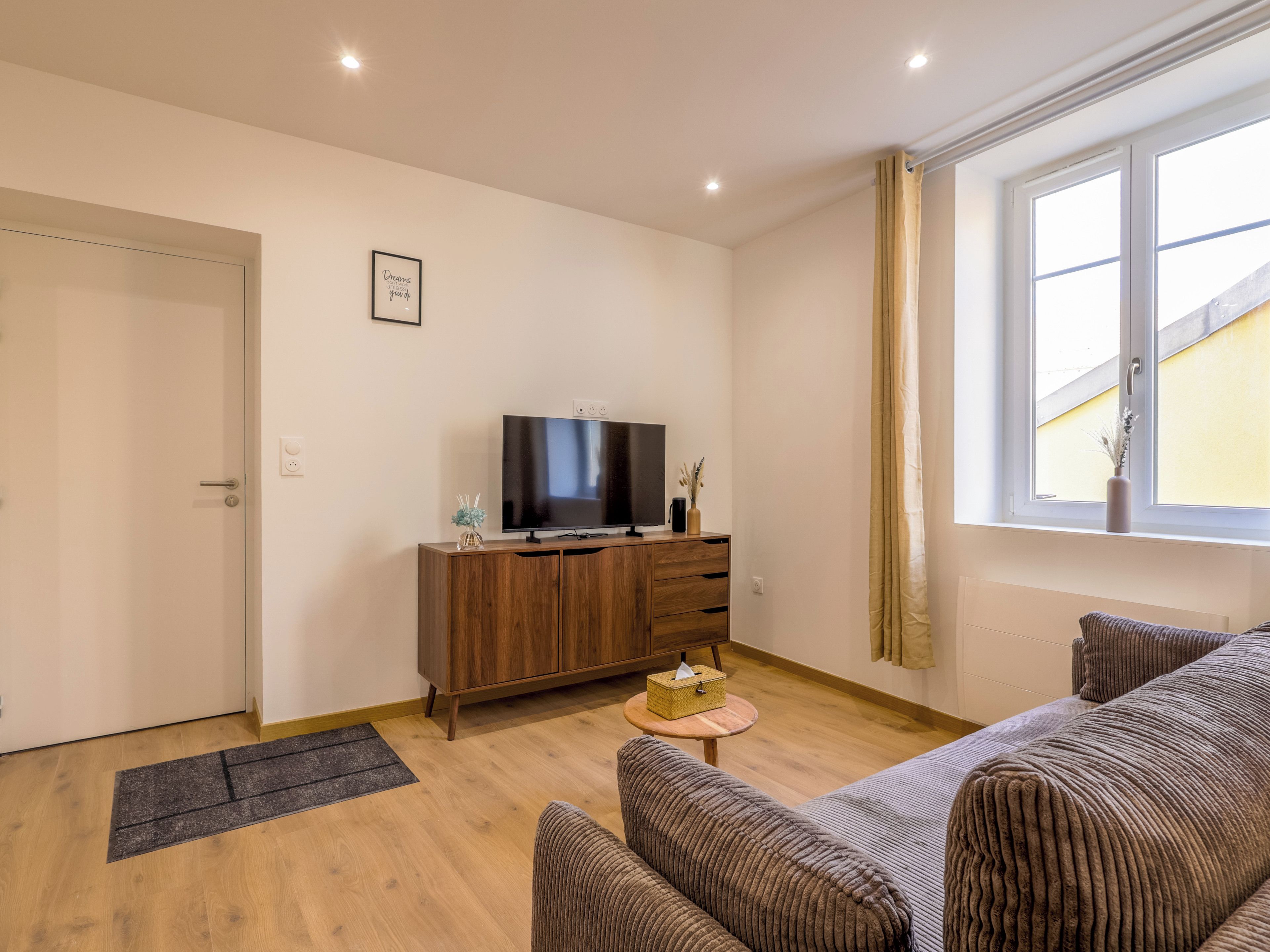 Een woonkamer met een bank, een televisie op een houten kast, een bijzettafeltje met een vaas, en een raam met gordijnen.