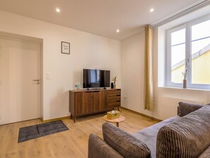 A living room with a sofa, a television on a wooden cabinet, a small table with a vase, and a window with curtains. - Écume du Lac - Gérardmer, Vosges (Gérardmer)