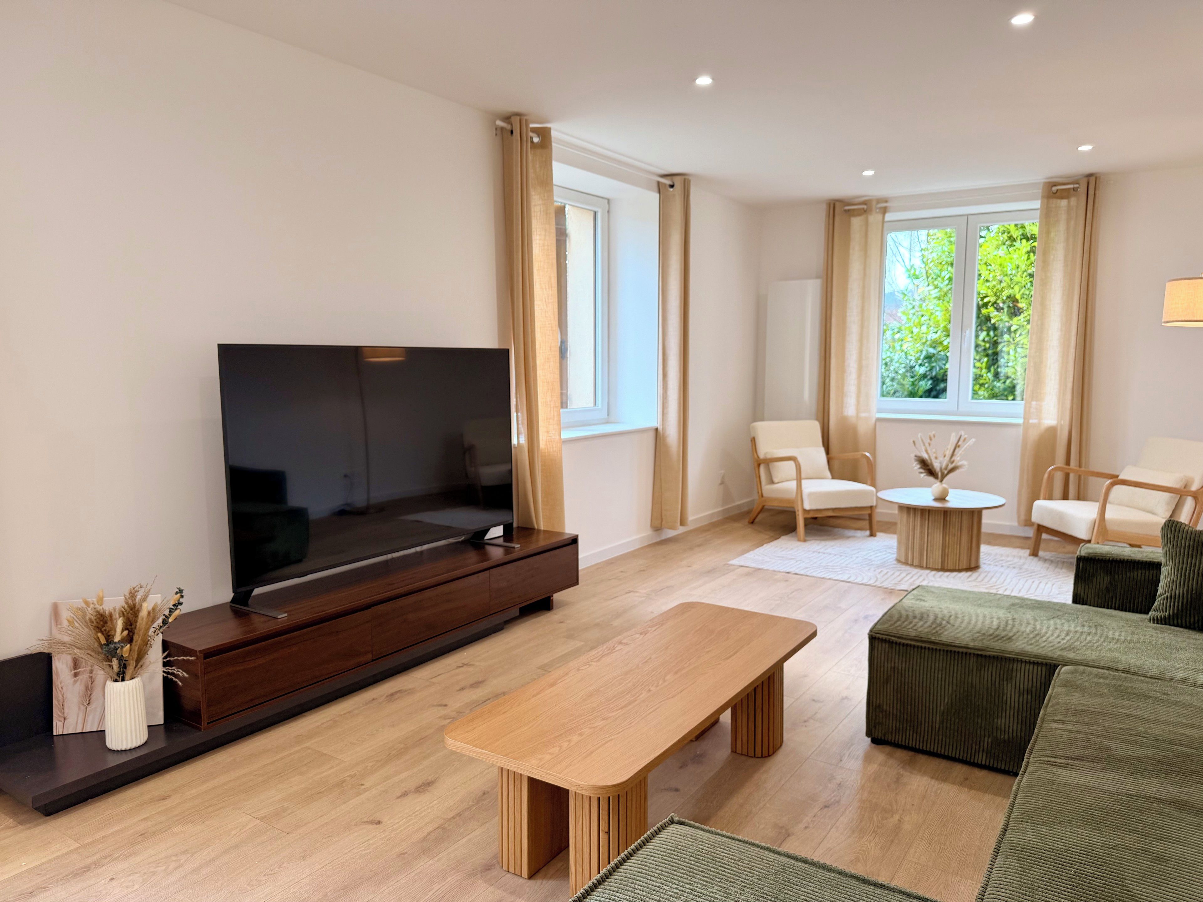 A modern living room with a flat-screen TV, wooden furniture, and large windows.
