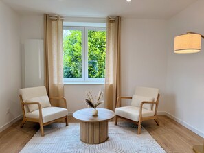 Ein Zimmer mit zwei Sesseln, einem runden Tisch, einer Vase mit Trockenpflanzen, einem Fenster mit Vorhängen und einer Hängelampe.