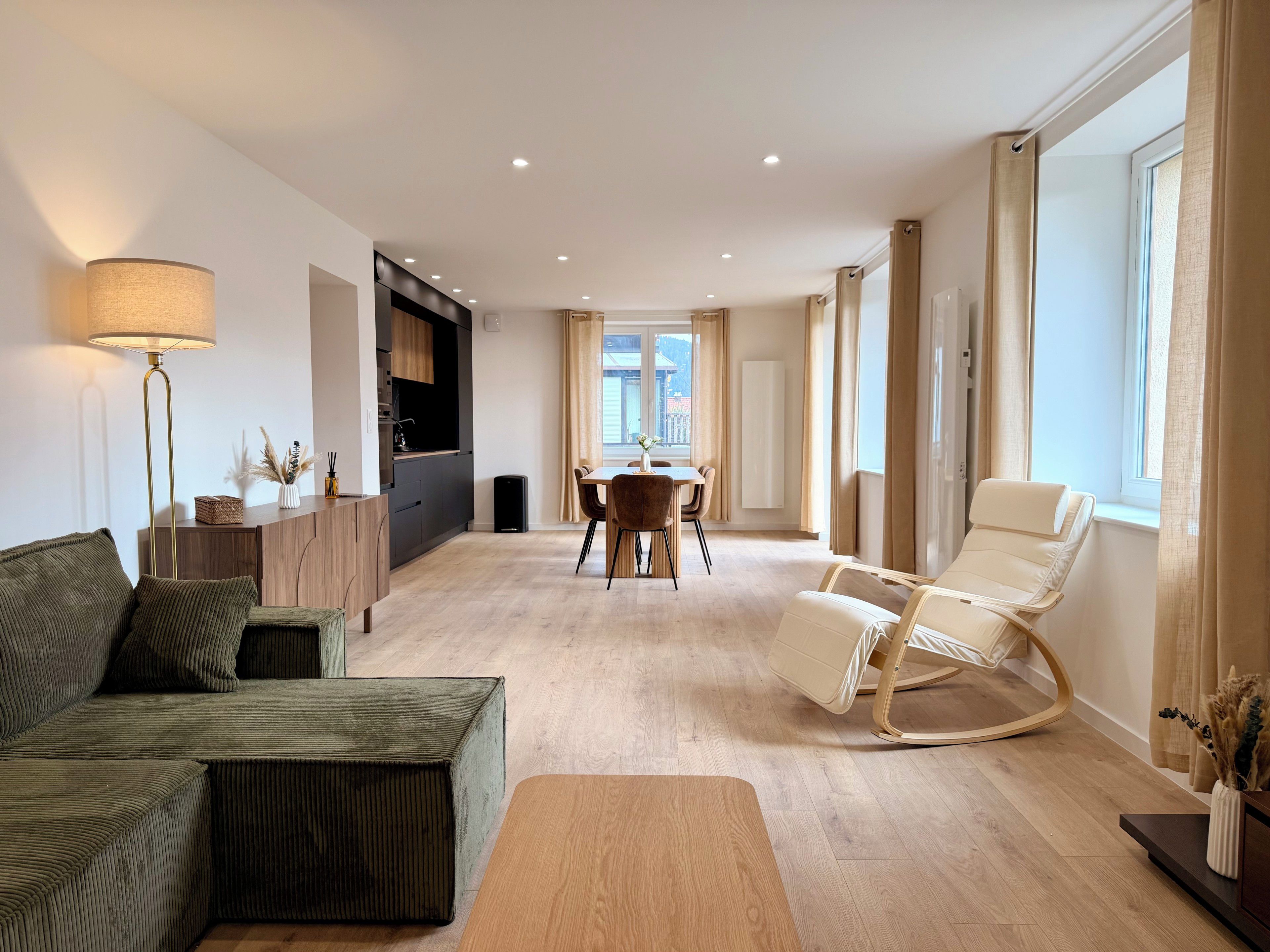 A modern living room with a sofa, a coffee table, a dining area with chairs, and a large window with curtains.