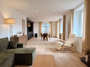 Ein modernes Wohnzimmer mit einer Couch, einem Couchtisch, einem Essbereich mit Stühlen und einem großen Fenster mit Vorhängen.